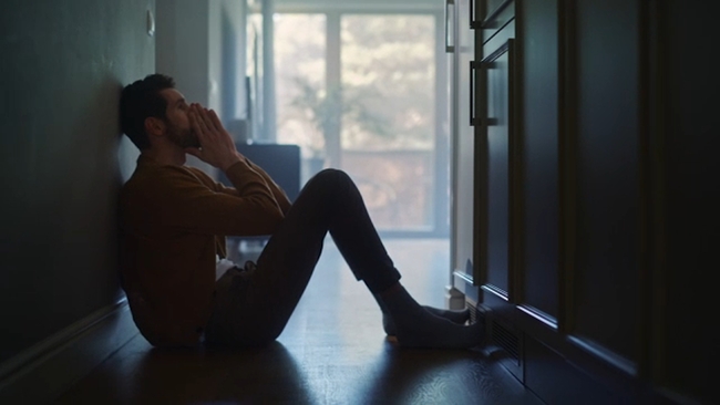 Man sitting alone on floor in a dim hallway, appearing distressed and isolated