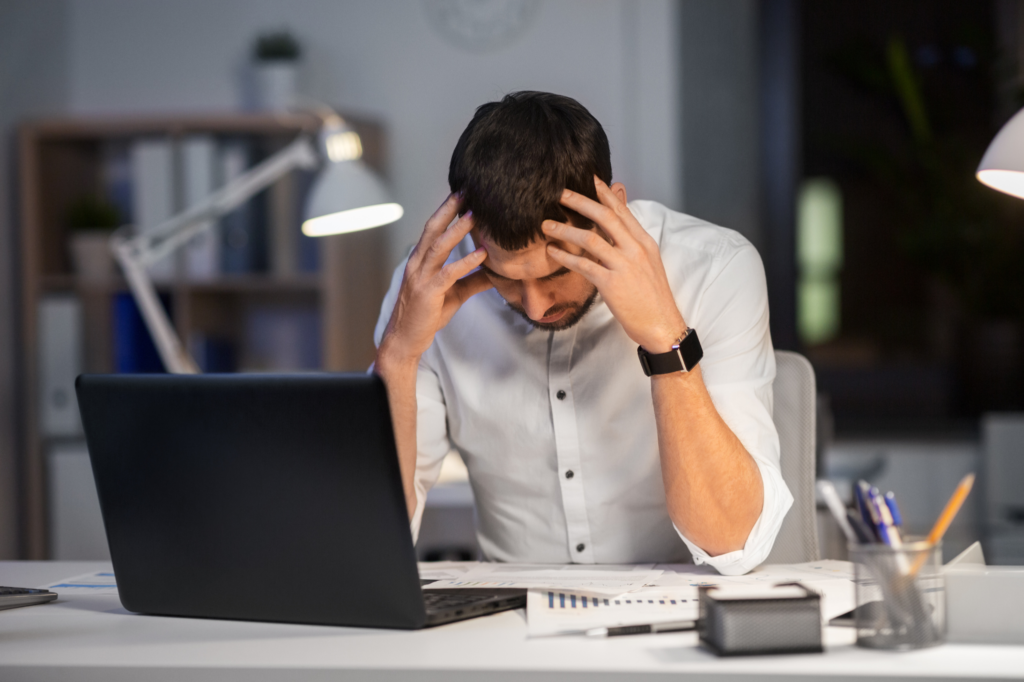 Stressed office worker sitting at a desk with a laptop, holding his head in frustration while reviewing paperwork in a dimly lit workspace.