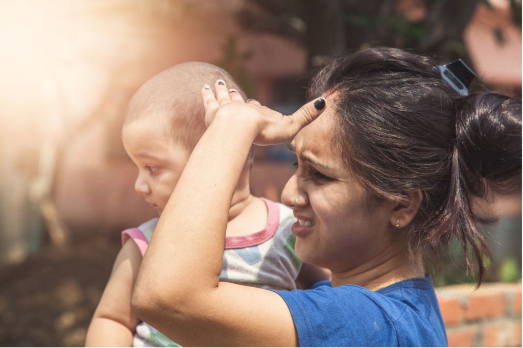 Mother holding a young child outdoors under bright sunlight while shielding her eyes with her hand, illustrating the effects of extreme heat and sun exposure.